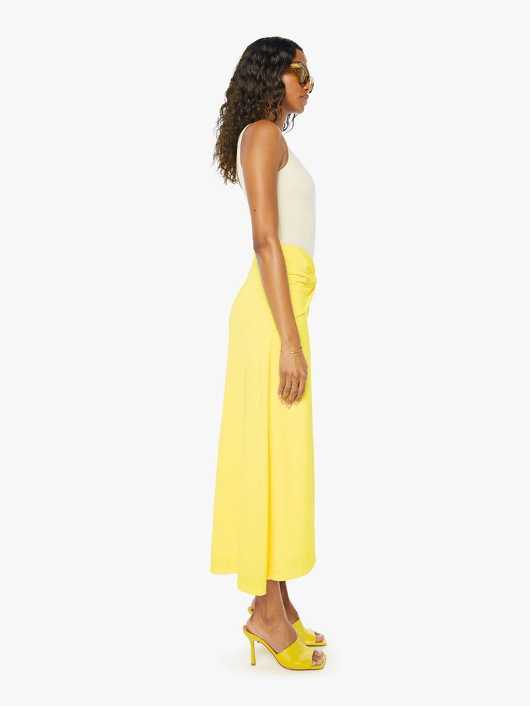 A side view of a woman wearing a bright yellow skirt with a flowy fit, featuring a front knotted wrap detail, paired with a white tank top.