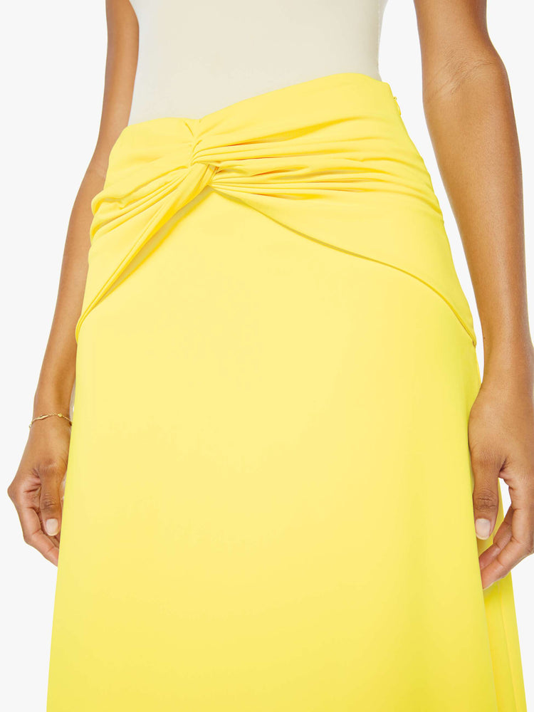 A front close up view of a woman wearing a bright yellow skirt with a flowy fit, featuring a front knotted wrap detail, paired with a white tank top.