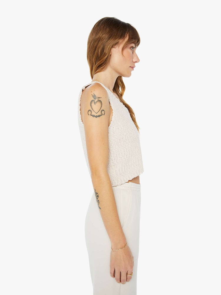 Side view of a woman wearing a knit tank in an off white hue, featuring a cropped body.