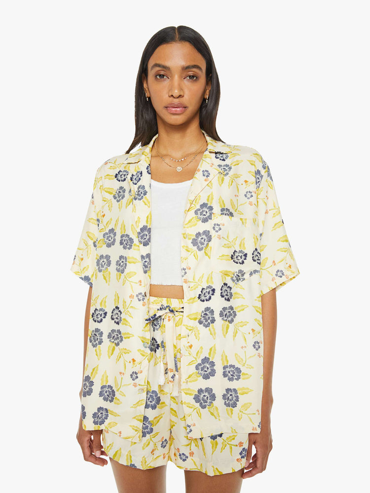 Front view of a woman in a unisex oversized shirt in a pale yellow, indigo and white floral print, and features a V-neck, drop shoulders, elbow-length sleeves, buttons down the front and a long, thigh-grazing hem.