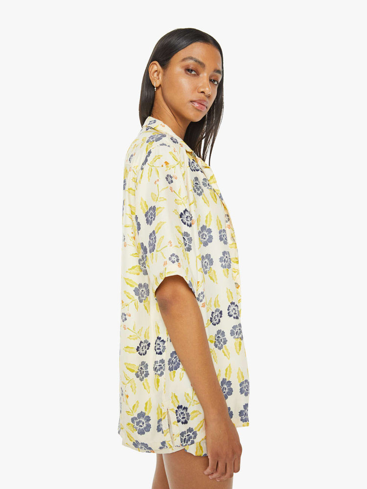 Side view of a woman in a unisex oversized shirt in a pale yellow, indigo and white floral print, and features a V-neck, drop shoulders, elbow-length sleeves, buttons down the front and a long, thigh-grazing hem.