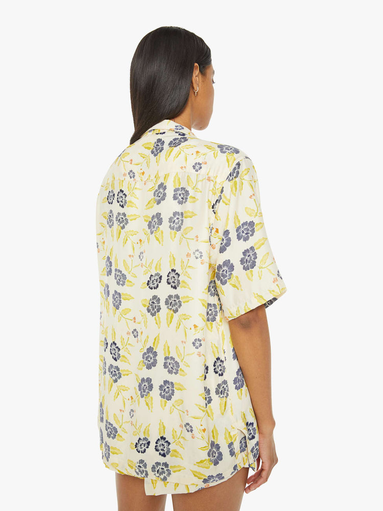 Back view of a woman in a unisex oversized shirt in a pale yellow, indigo and white floral print, and features a V-neck, drop shoulders, elbow-length sleeves, buttons down the front and a long, thigh-grazing hem.