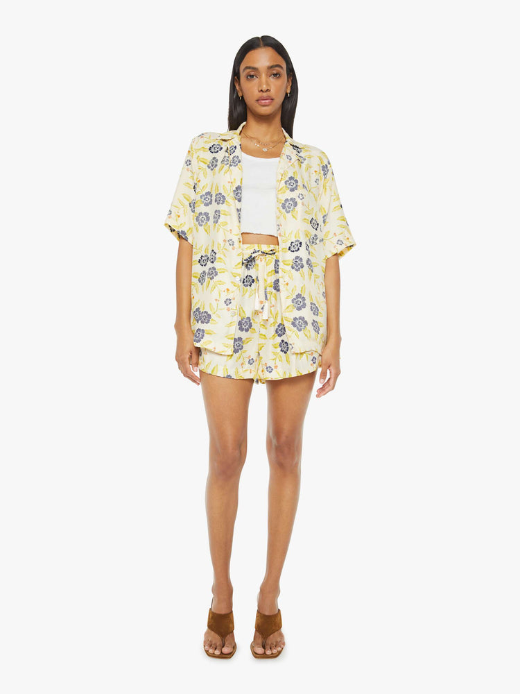 Full body view of a woman in a unisex oversized shirt in a pale yellow, indigo and white floral print, and features a V-neck, drop shoulders, elbow-length sleeves, buttons down the front and a long, thigh-grazing hem.