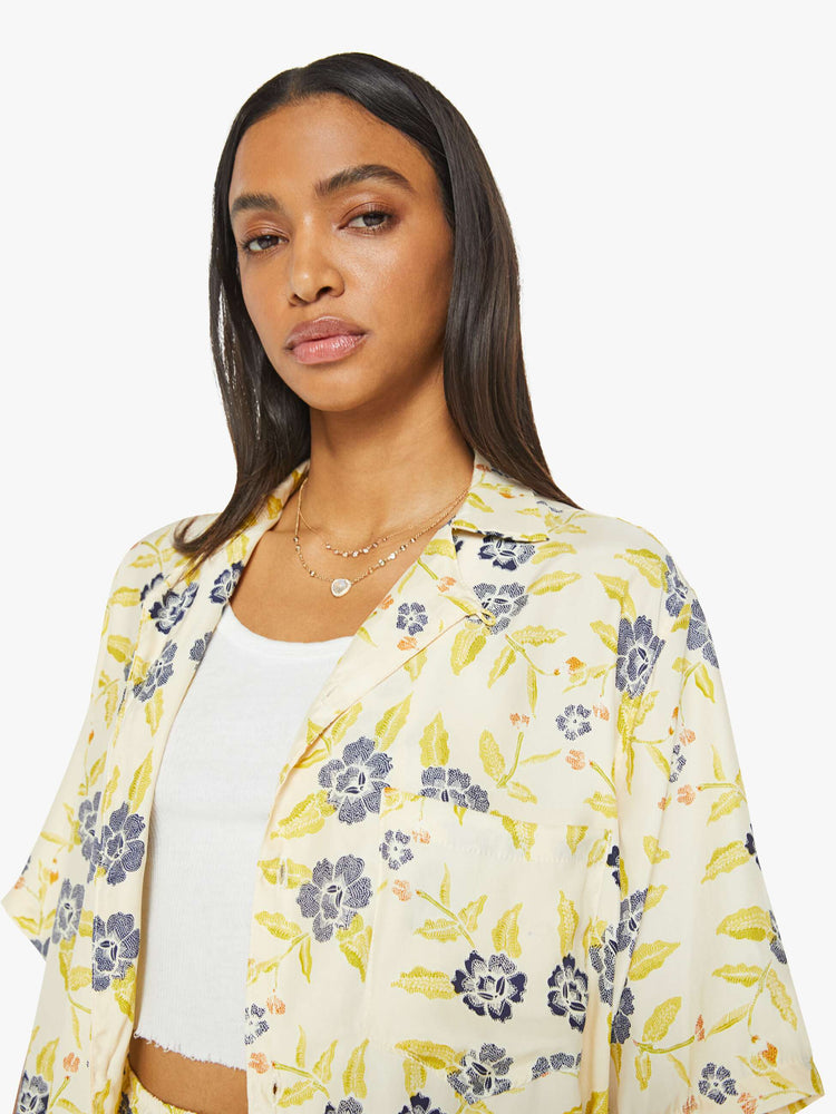 Close up view of a woman in a unisex oversized shirt in a pale yellow, indigo and white floral print, and features a V-neck, drop shoulders, elbow-length sleeves, buttons down the front and a long, thigh-grazing hem.