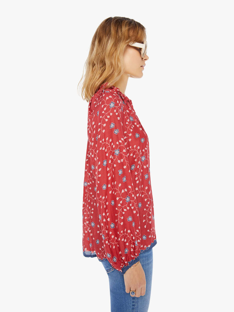 Side view of a woman wearing a red blouse with blue floral print, long sleeves, and tassel closure at the neckline