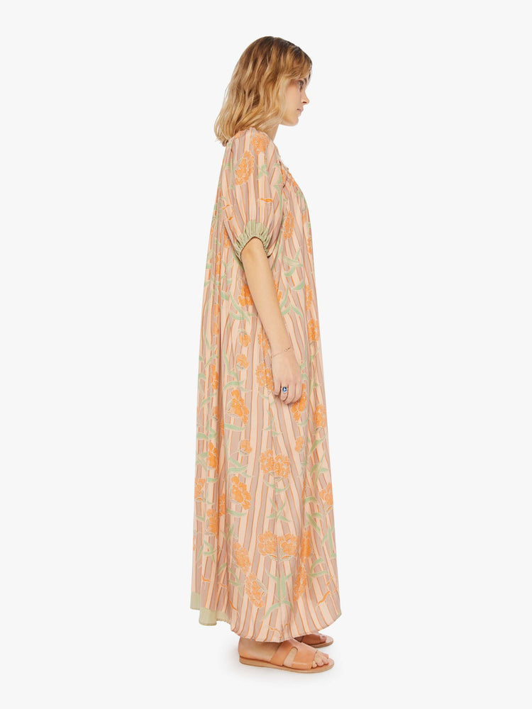 side  view of a woman in a cream maxi dress with stripes and a warm-toned floral print, designed with a scoop neck, elbow-length sleeves and an A-line cut for a loose, breezy feel.