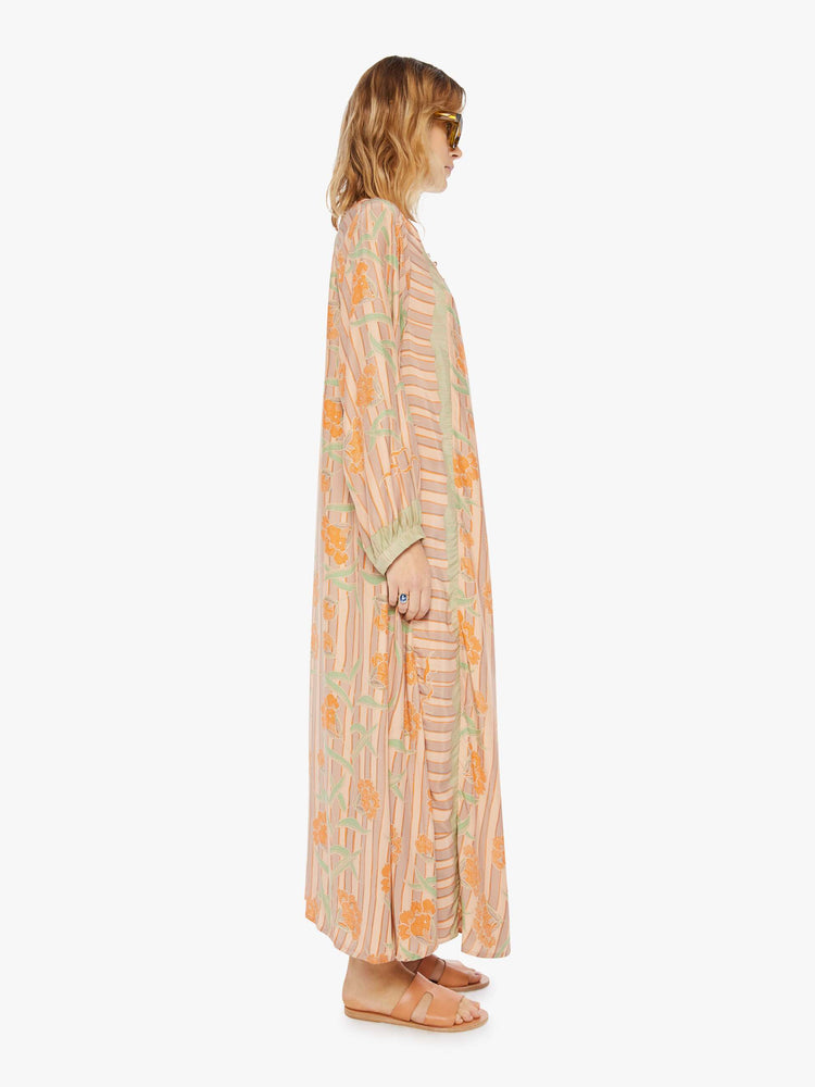 side view of a woman in a cream dress with stripes and a warm-toned floral print. This maxi dress is designed with voluminous sleeves and has an A-line cut for a loose, breezy feel.