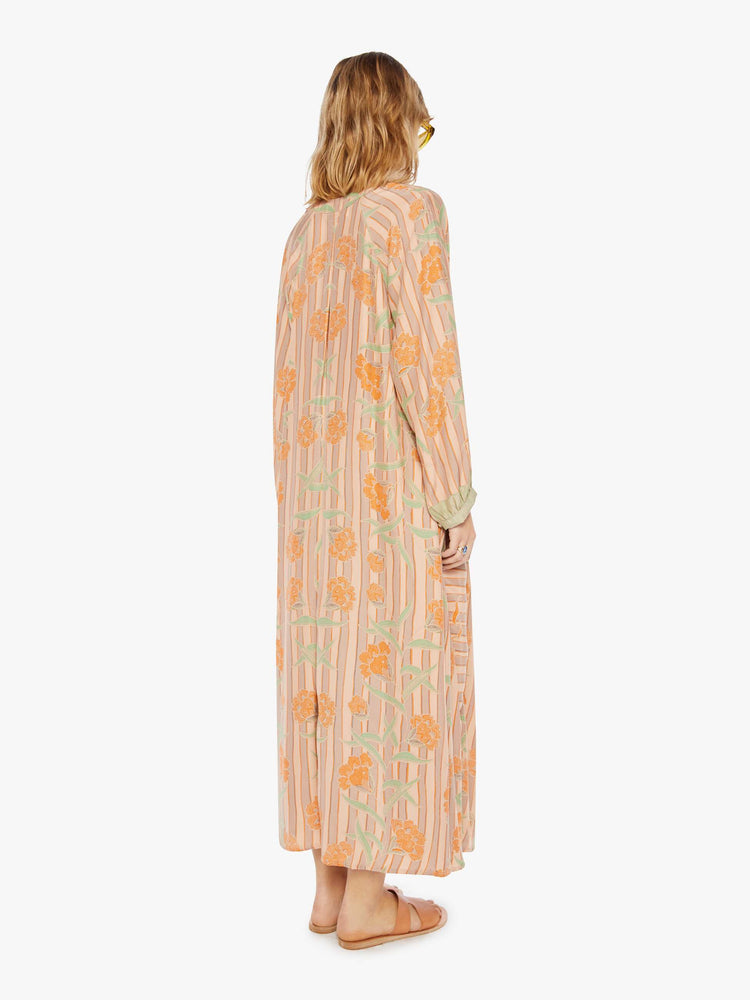 back view of a woman in a cream dress with stripes and a warm-toned floral print. This maxi dress is designed with voluminous sleeves and has an A-line cut for a loose, breezy feel.