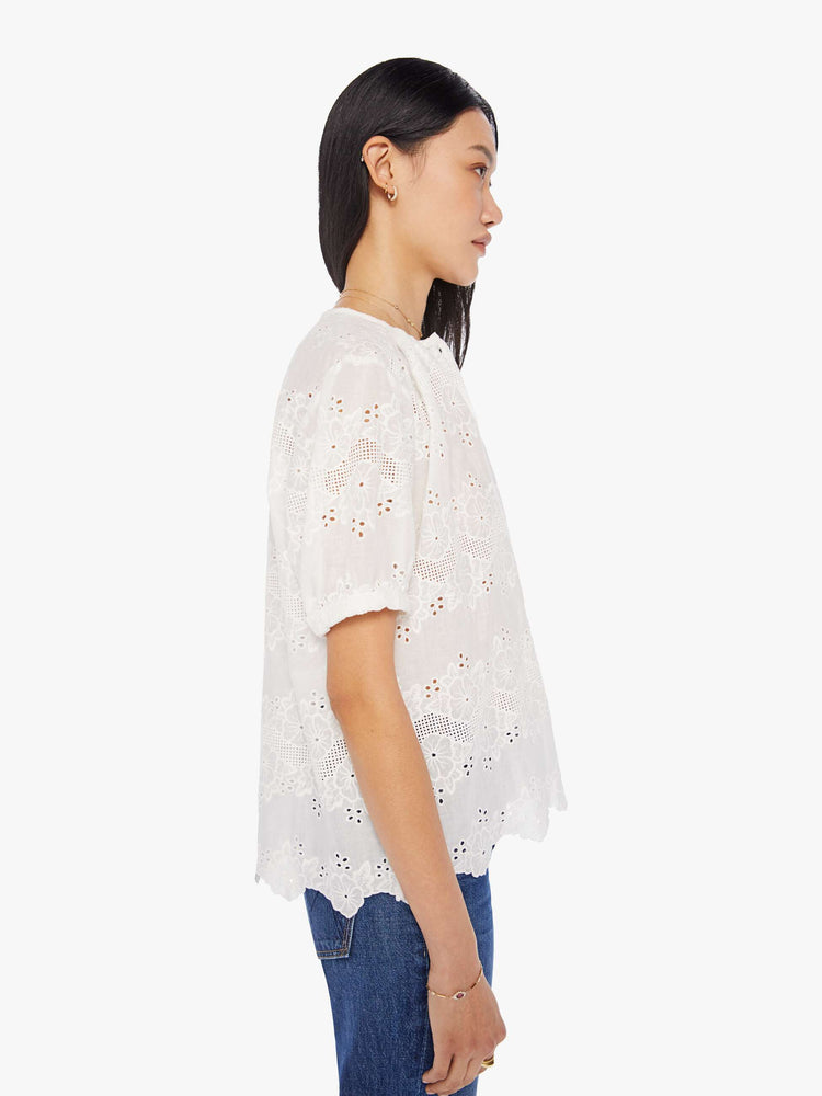 side view of a woman in a cream top with an eyelet floral print and a scalloped hem, designed with a deep V-neck, buttons down the front, short balloon sleeves and ruffles throughout for a loose, flowy fit.