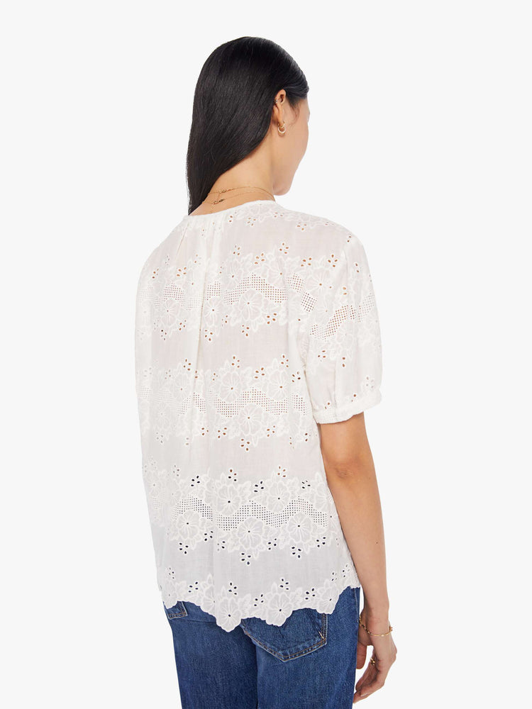 back view of a woman in a cream top with an eyelet floral print and a scalloped hem, designed with a deep V-neck, buttons down the front, short balloon sleeves and ruffles throughout for a loose, flowy fit.