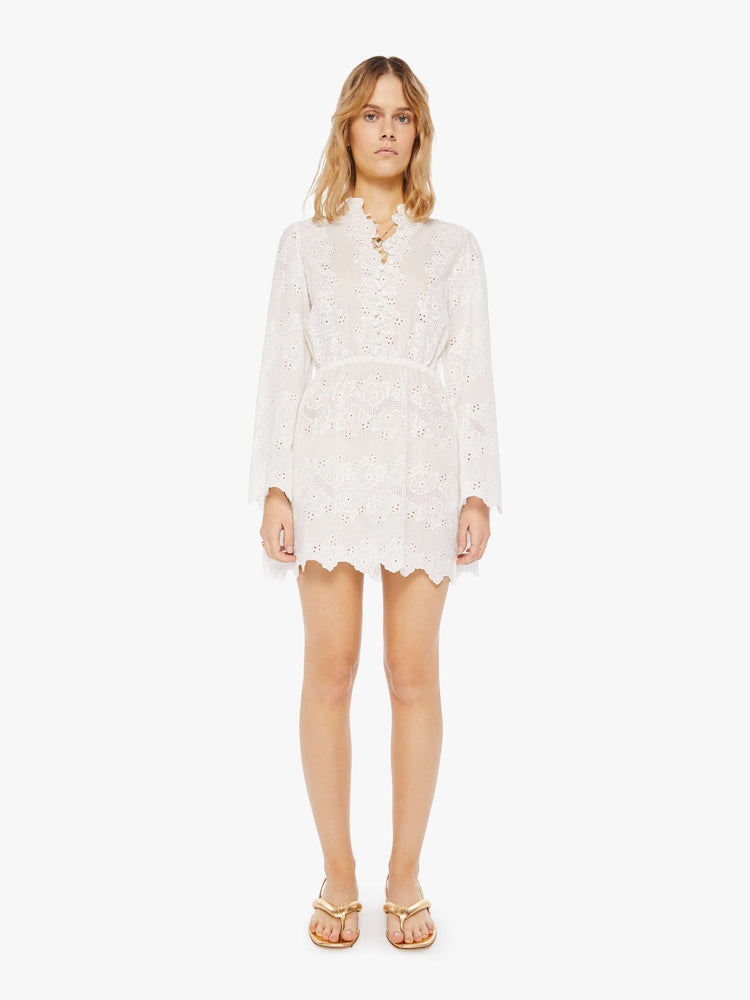Front view of a woman in a cream dress with openwork details. The mini dress features a crewneck with a short collar, long flowy sleeves, a fitted waist and zig-zag hems.
