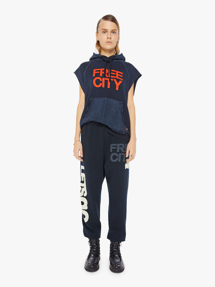 A front full body view of a woman wearing an oversized navy sweatshirt hoodie with raw cutoff sleeves and a large front graphic reading "FREE CITY", paired with a dark navy sweatpant.