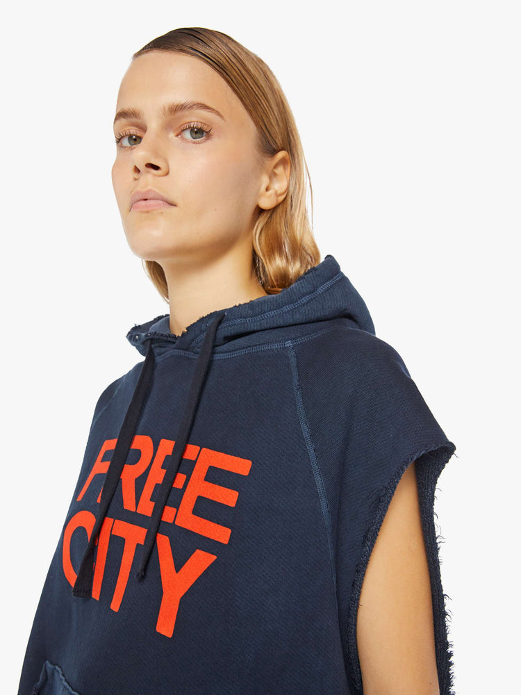 A front close up view of a woman wearing an oversized navy sweatshirt hoodie with raw cutoff sleeves and a large front graphic reading "FREE CITY".