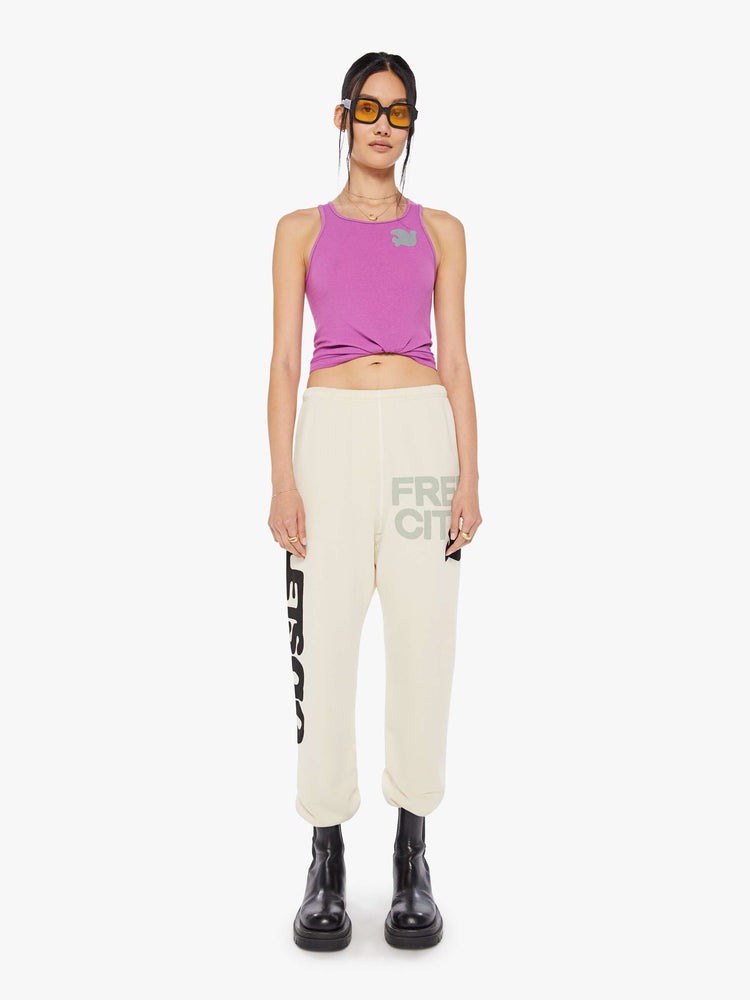 A front view of a woman wearing a bright purple ribbed tank top with a bird graphic on the chest, paired with an off white sweatpants.