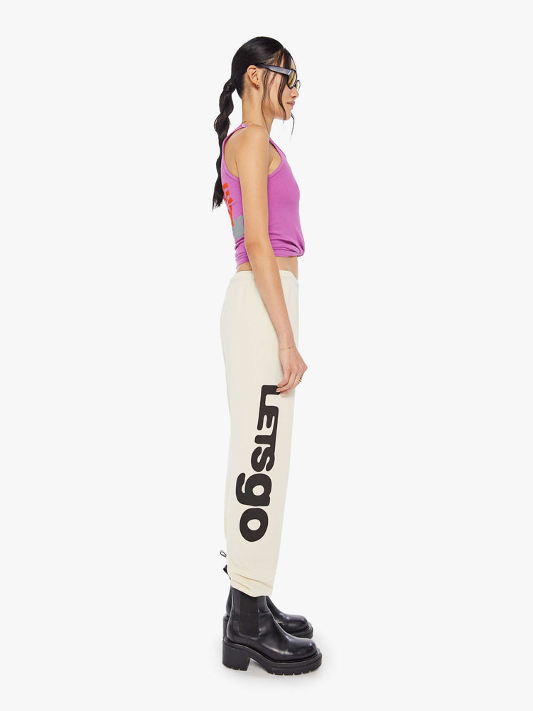 A side view of a woman wearing a bright purple ribbed tank top with a bird graphic on the chest, paired with an off white sweatpants.