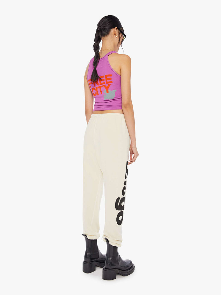 A back view of a woman wearing a bright purple ribbed tank top with a bird graphic on the chest, paired with an off white sweatpants.