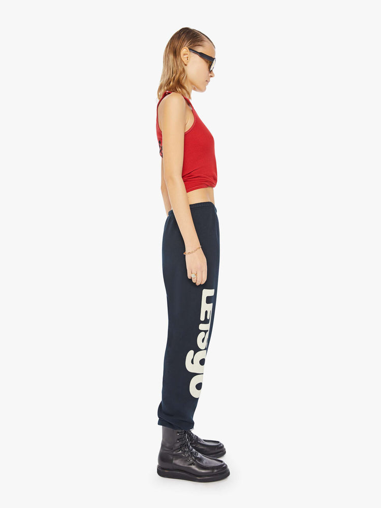 A side view of a woman wearing a bright red ribbed tank top with a bird graphic on the chest, paired with a dark navy sweatpants.