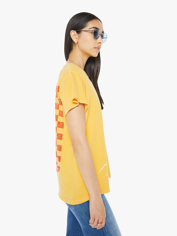 Side view of a woman tee in golden-yellow, the tee honors the Rolling Stones with the band's iconic tongue-and-lips logo on the front and a checker-print graphic on the back.