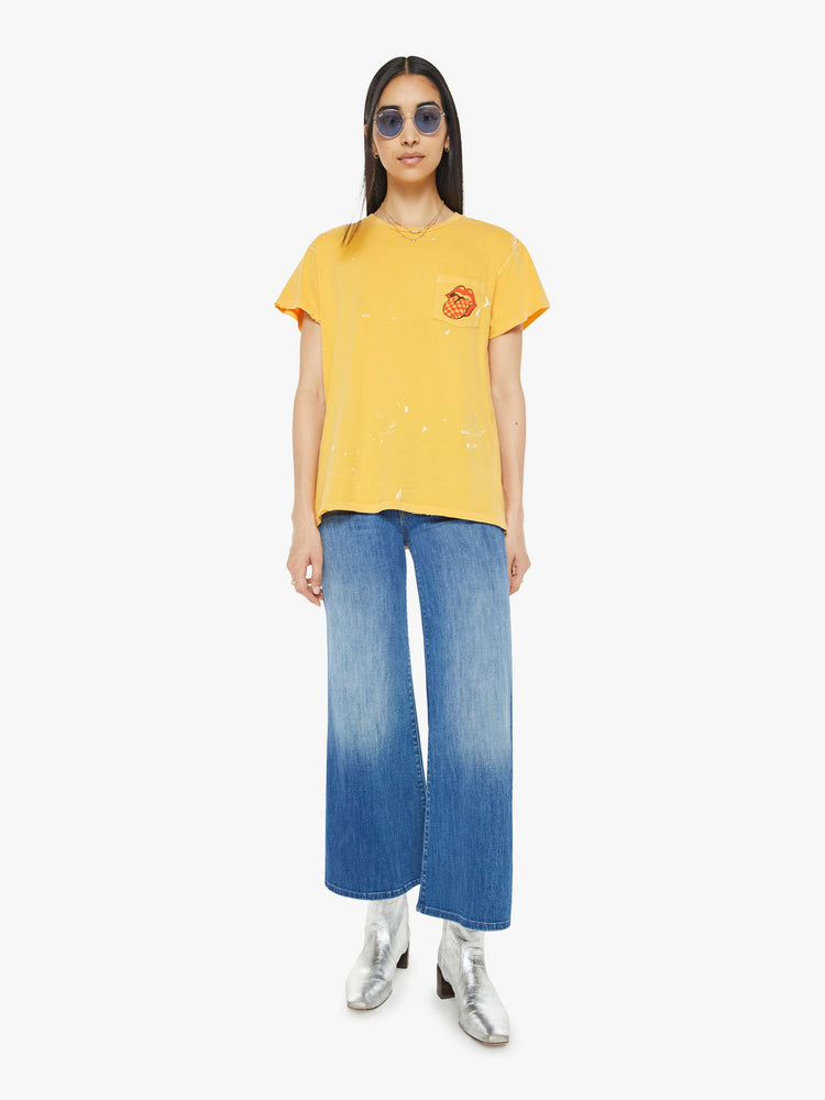 Full body view of a woman tee in golden-yellow, the tee honors the Rolling Stones with the band's iconic tongue-and-lips logo on the front and a checker-print graphic on the back.