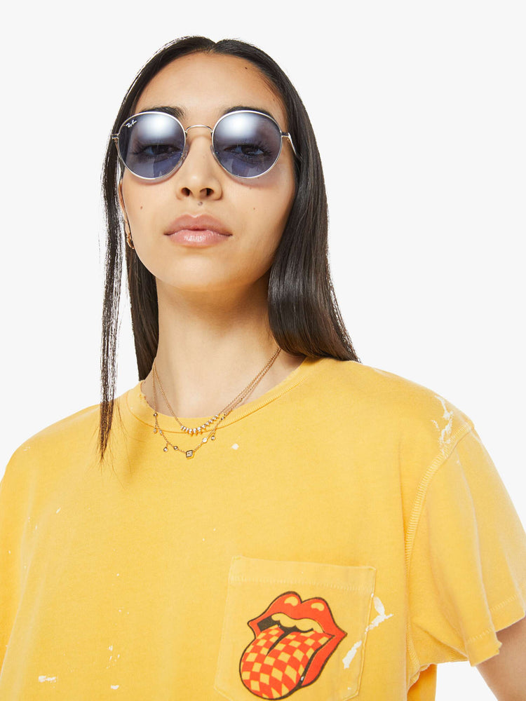 Close up view of a woman tee in golden-yellow, the tee honors the Rolling Stones with the band's iconic tongue-and-lips logo on the front and a checker-print graphic on the back.