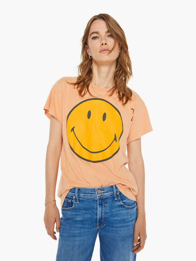 Front view of a woman peach tee with  a smiley face graphic on the front and text on the back. 