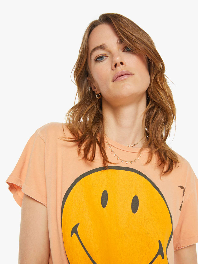 Close up view of a woman peach tee with a smiley face graphic on the front and text on the back.