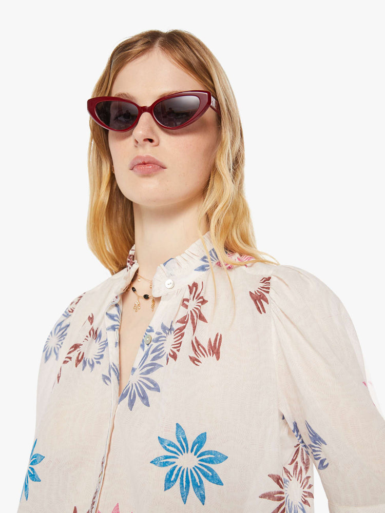 Close up view of a woman in a cream with a graphic floral print button down shirt with a stacked collar, puffed shoulders, elbow-length sleeves.