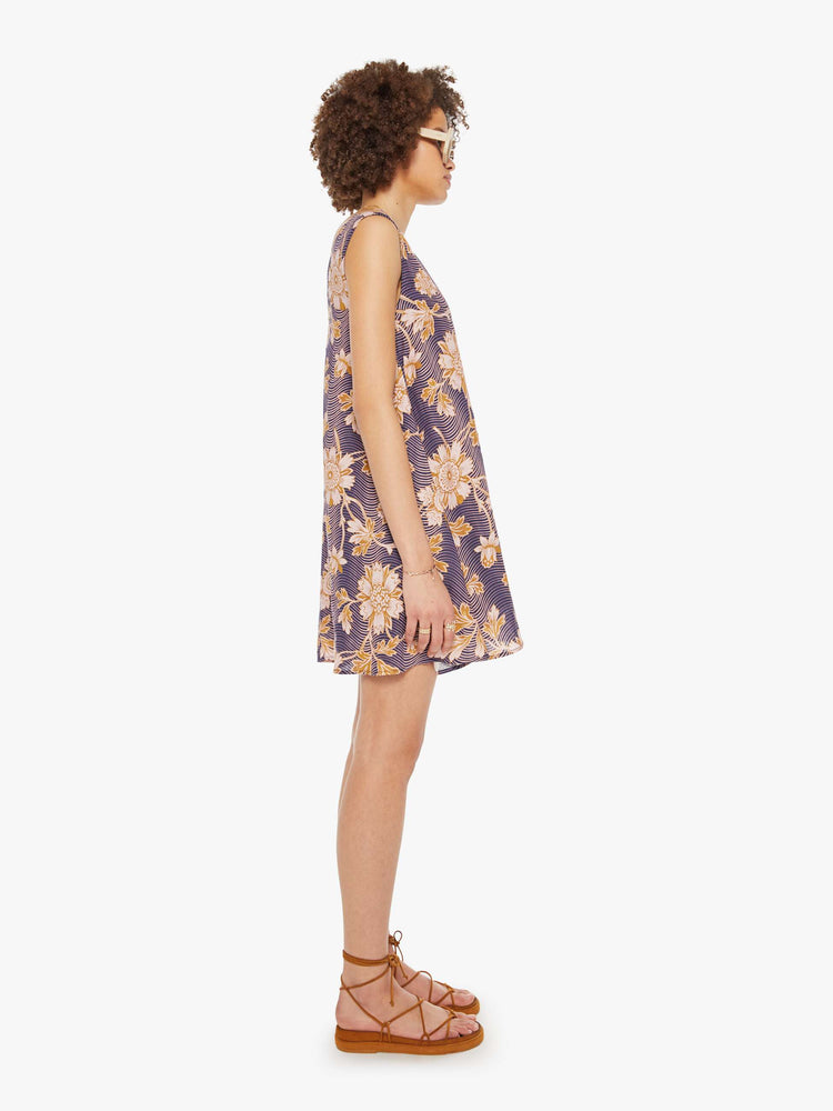 Side view of a woman in a purple with graphic floral print dress with a V-neck, wide straps and short fit.