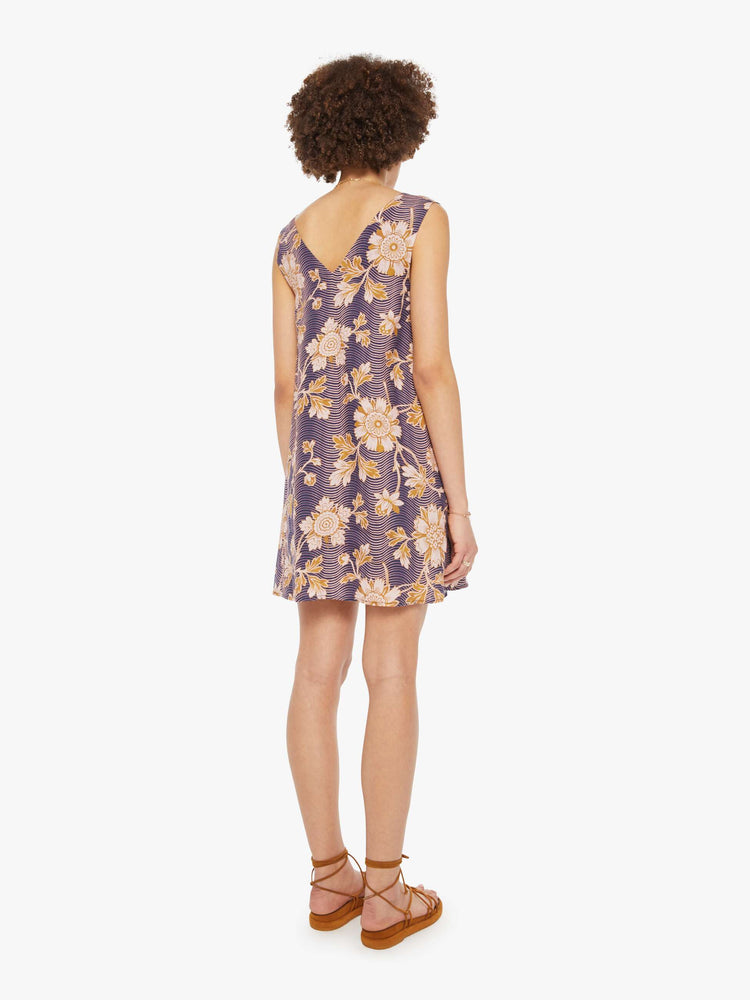 Back view of a woman in a purple with graphic floral print dress with a V-neck, wide straps and short fit.