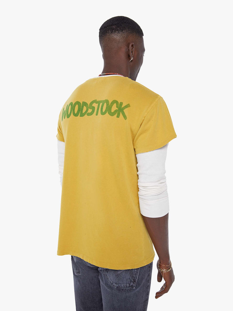 MEN back view in a  mustard yellow tee that pays homage to the legendary Woodstock festival with a green graphic on the front.