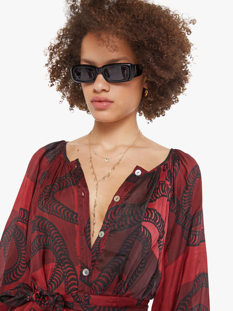 Close up view of a woman in a oxblood with a black snake graphic dress with a V-neck that buttons down the front, long balloon sleeves and ruffles throughout for a loose, flowy fit.