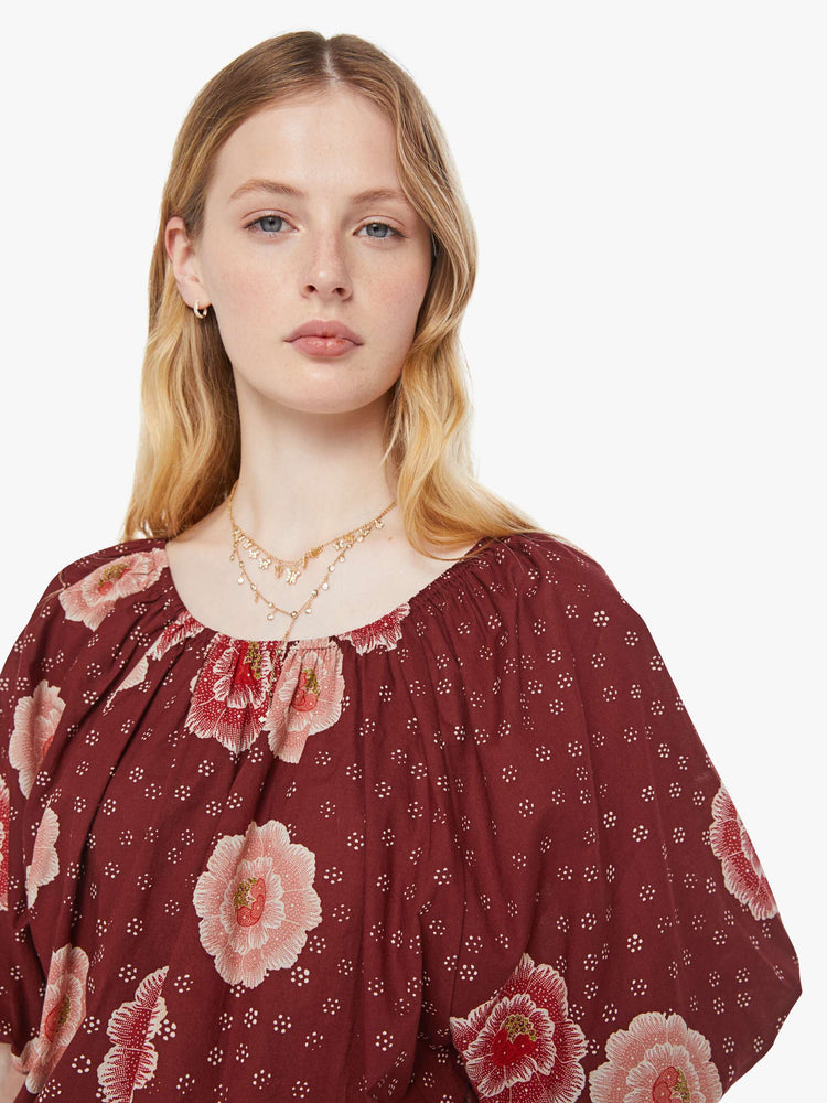 Close up view of a woman in a maroon with a pink poppy print shirt with elastic boat neck that can be worn off-the-shoulder, 3/4-length balloon sleeves and a flowy fit.