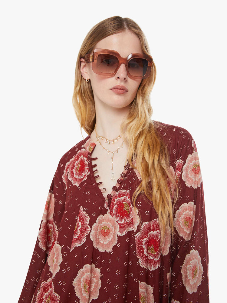 Close up view of a woman in a  maroon with a pink poppy print, this maxi dress is designed with voluminous sleeves and has an A-line cut for a loose, breezy feel.