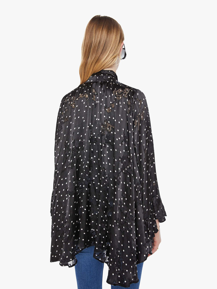 Back view of a woman in a black with white polka dots cape with a tied V-neck, open front, long billowy sleeves with ruffles at the wrists and a cropped hem.