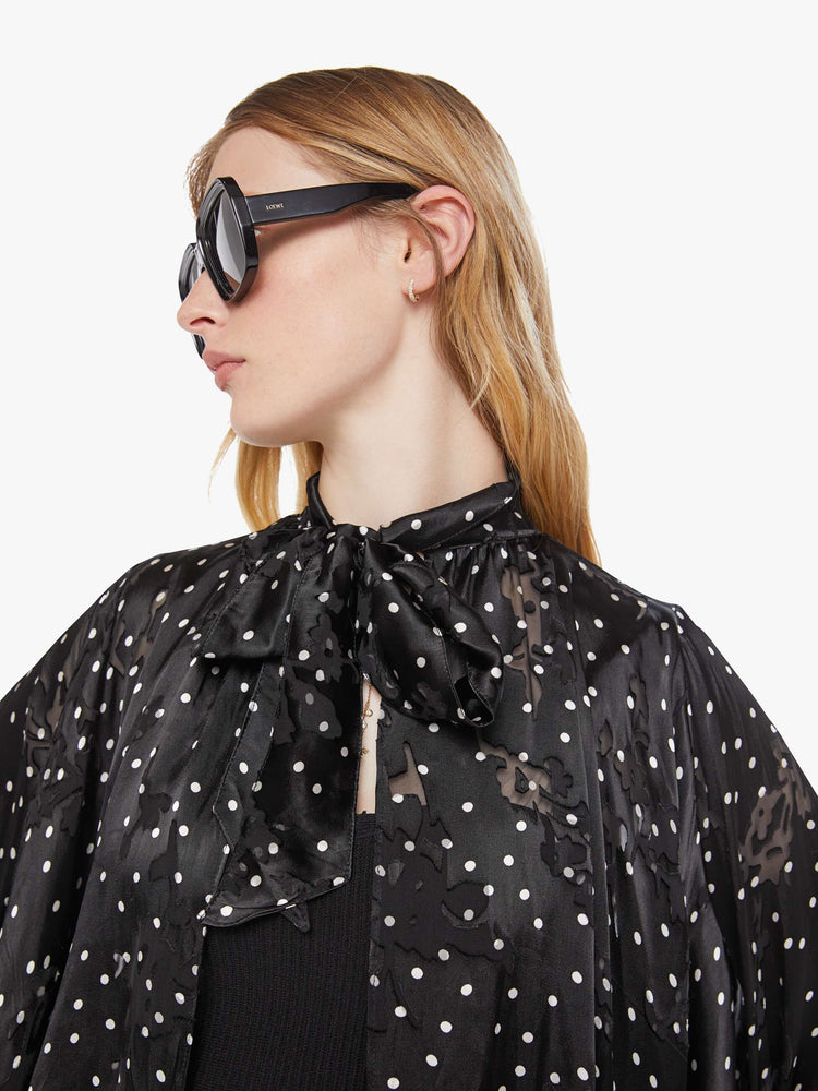 Close up view of a woman in a black with white polka dots cape with a tied V-neck, open front, long billowy sleeves with ruffles at the wrists and a cropped hem.