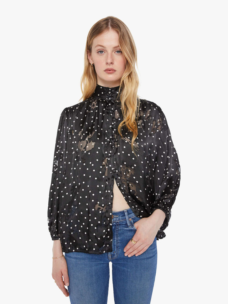 Front view of a woman in black with a white polka dot print top with a buttoned thick band collar, buttons down the front, balloon sleeves and a loose, flowy fit.