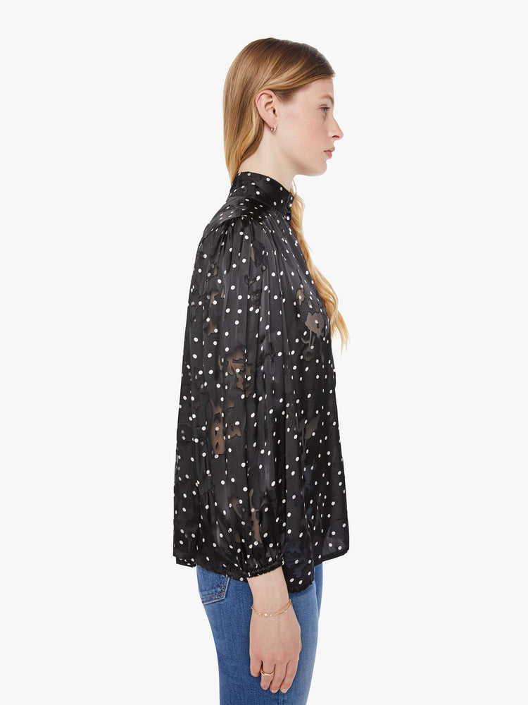 Side view of a woman in black with a white polka dot print top with a buttoned thick band collar, buttons down the front, balloon sleeves and a loose, flowy fit.