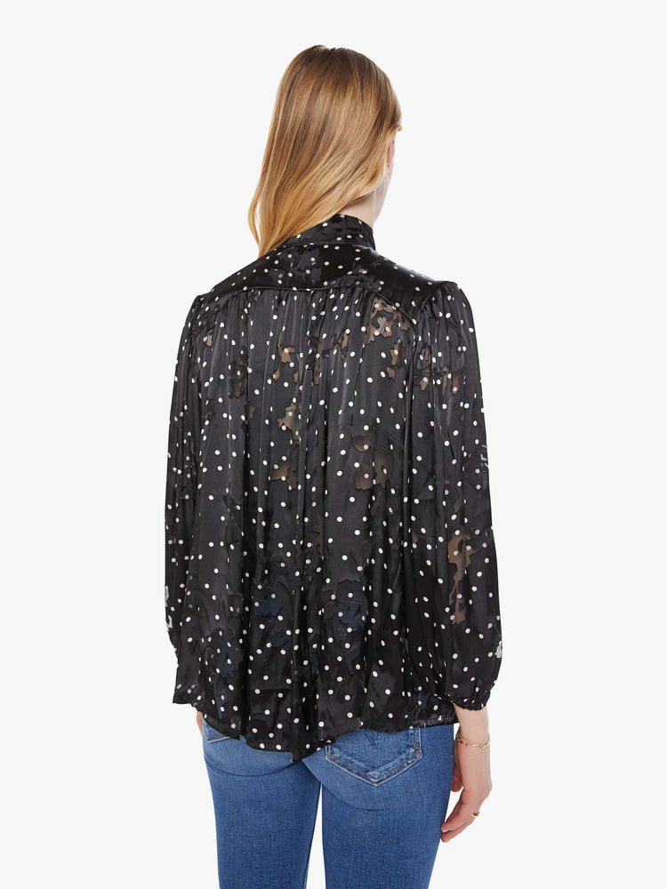 Back view of a woman in black with a white polka dot print top with a buttoned thick band collar, buttons down the front, balloon sleeves and a loose, flowy fit.