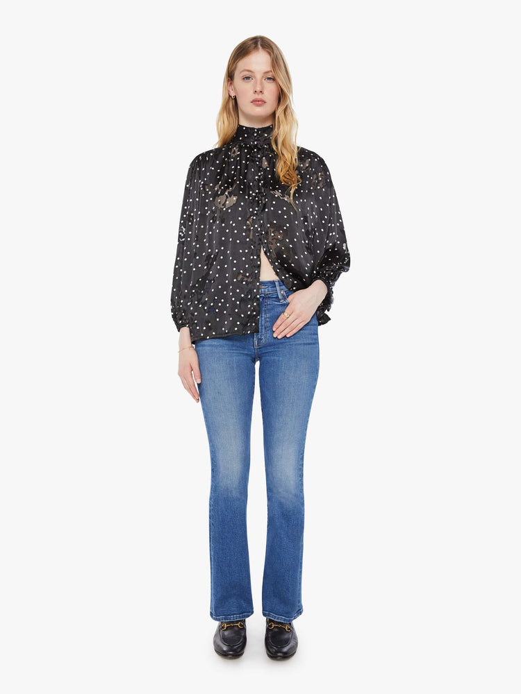 Full body view of a woman in black with a white polka dot print top with a buttoned thick band collar, buttons down the front, balloon sleeves and a loose, flowy fit.