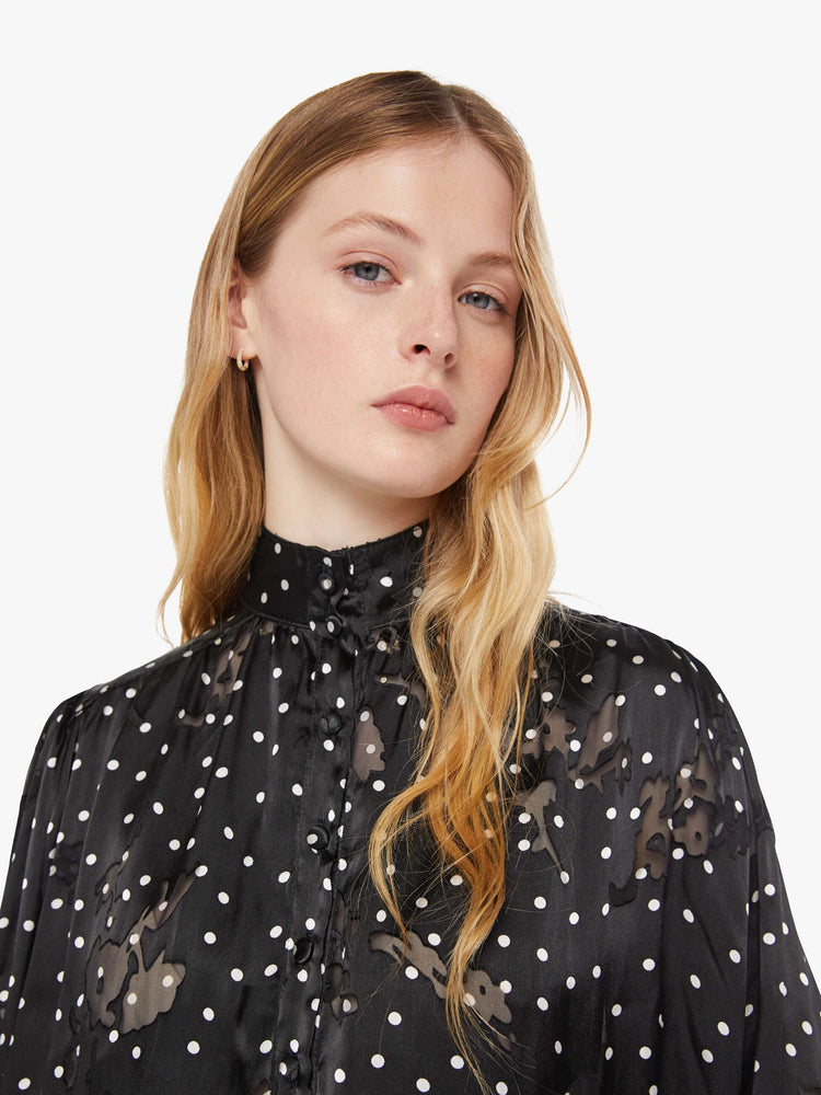 Close up view of a woman in black with a white polka dot print top with a buttoned thick band collar, buttons down the front, balloon sleeves and a loose, flowy fit.