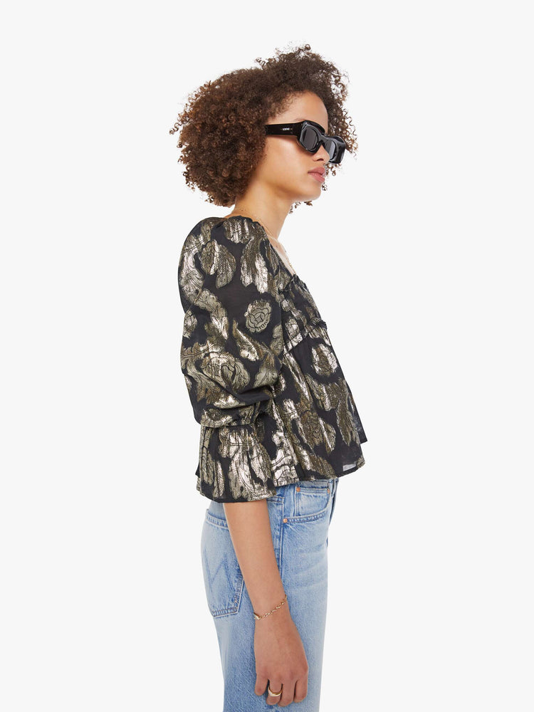 Side view of woman top in black with a metallic graphic print, and features a square neck, elbow-length balloon sleeves with a wide ruffled hem and a cropped, flowy fit.