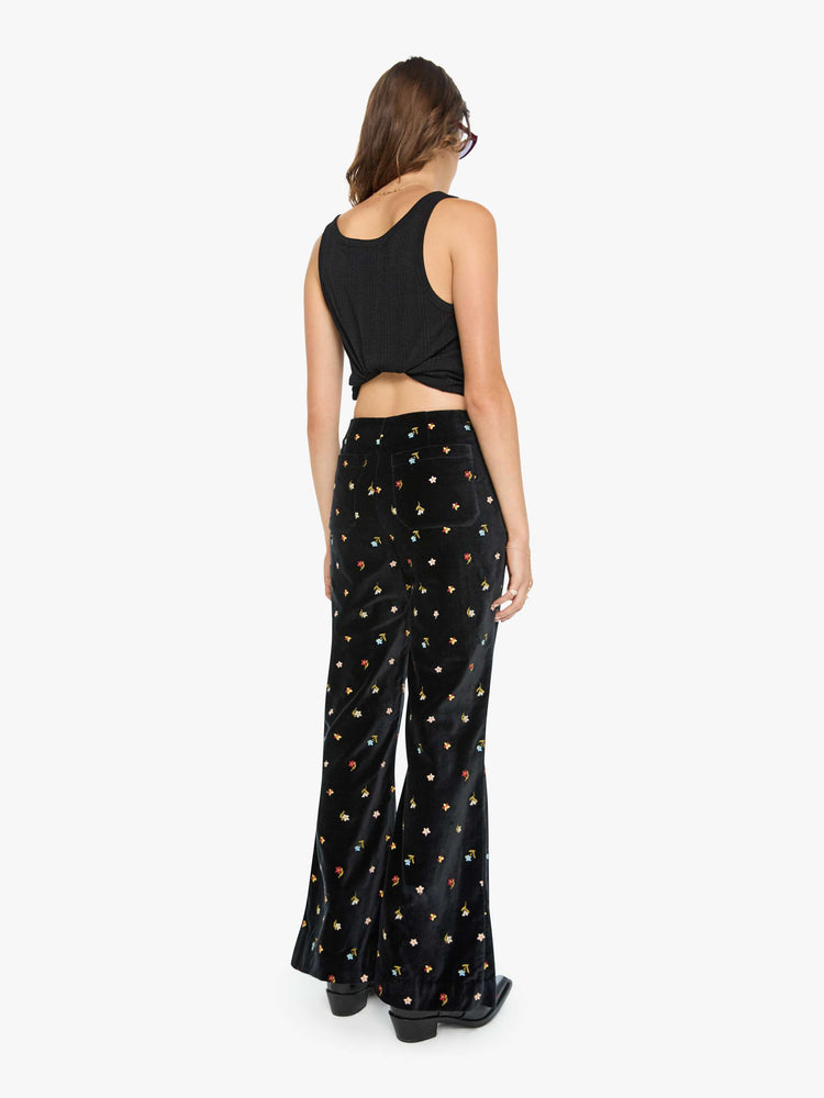 Back view of a woman in black with colorful floral details pants with a mid rise and a wide flared leg.