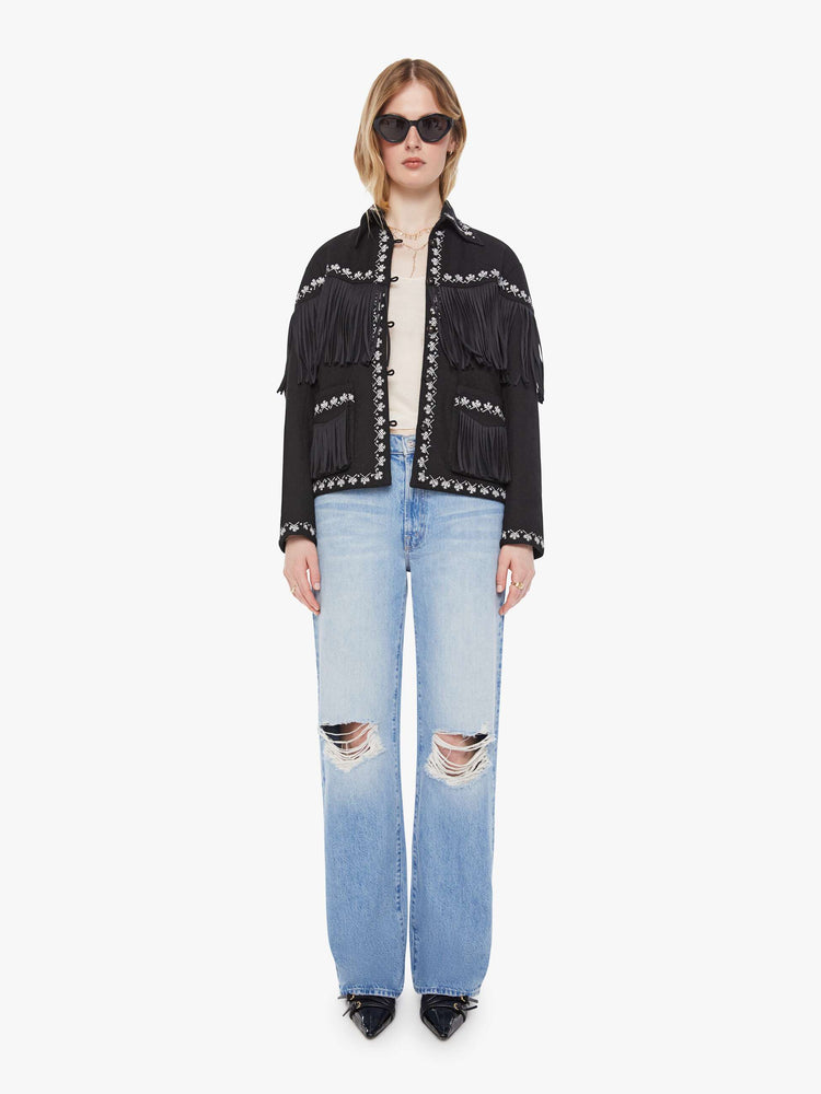 Full body view of a woman in a Jacket in a black with fringe details and white embroidery with dropped shoulders, front patch pockets, buttons down the front and a boxy fit.