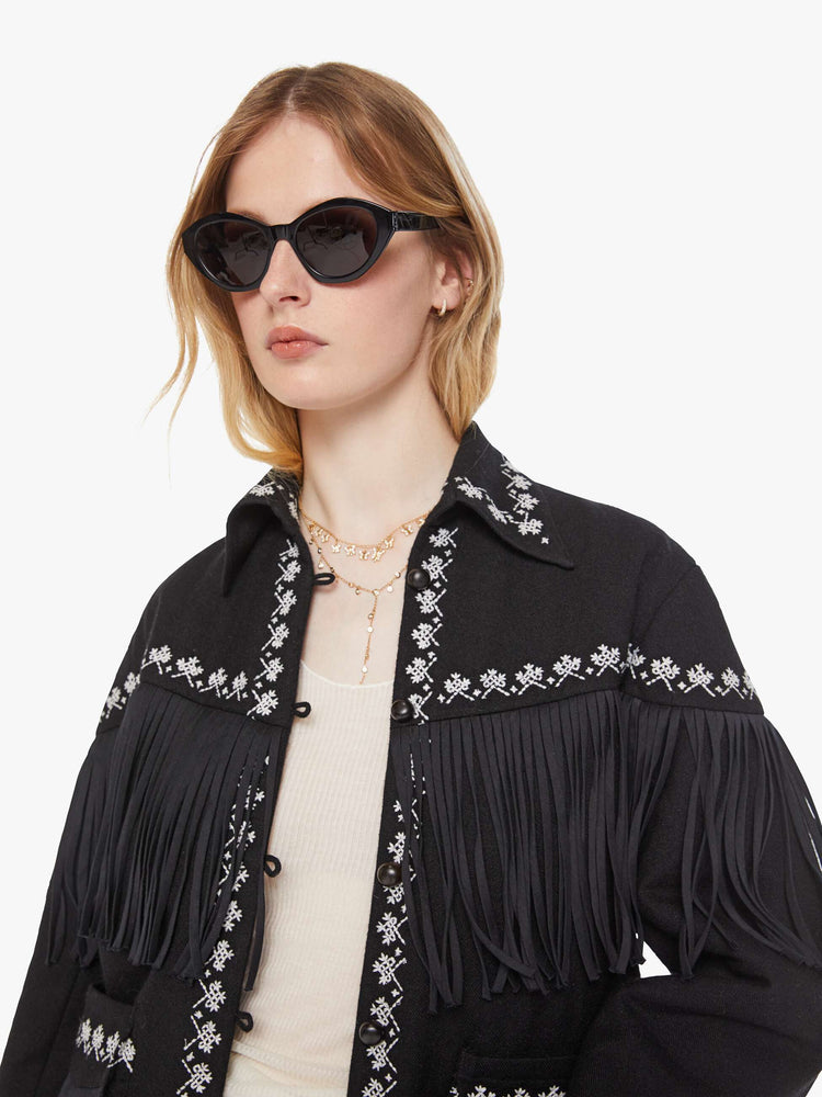 Close up view of a woman in a Jacket in a black with fringe details and white embroidery with dropped shoulders, front patch pockets, buttons down the front and a boxy fit.