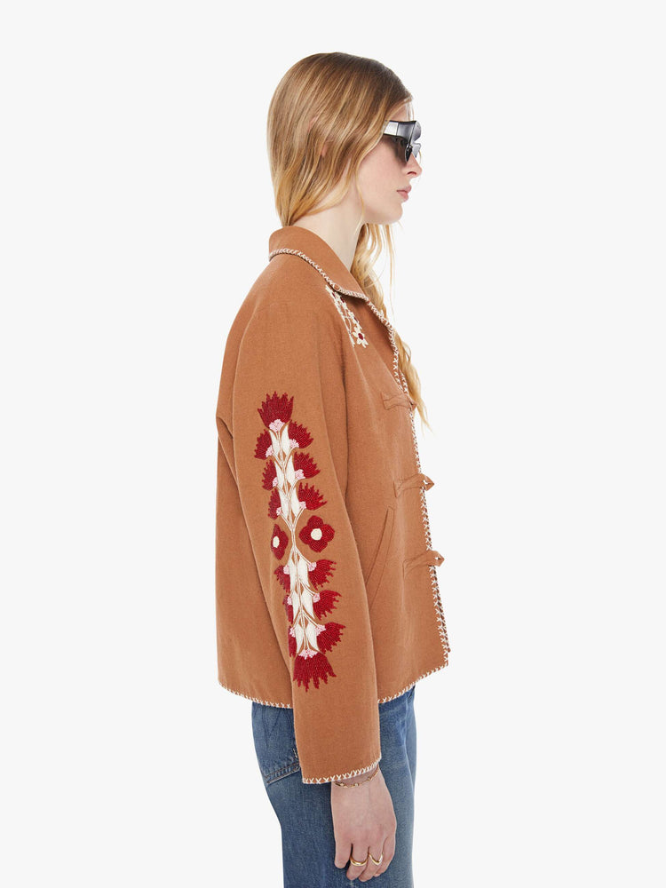 Side view of a woman in a  Western jacket in camel with embroidered motifs throughout. It has a notched collar, buttons down the front and a boxy fit.