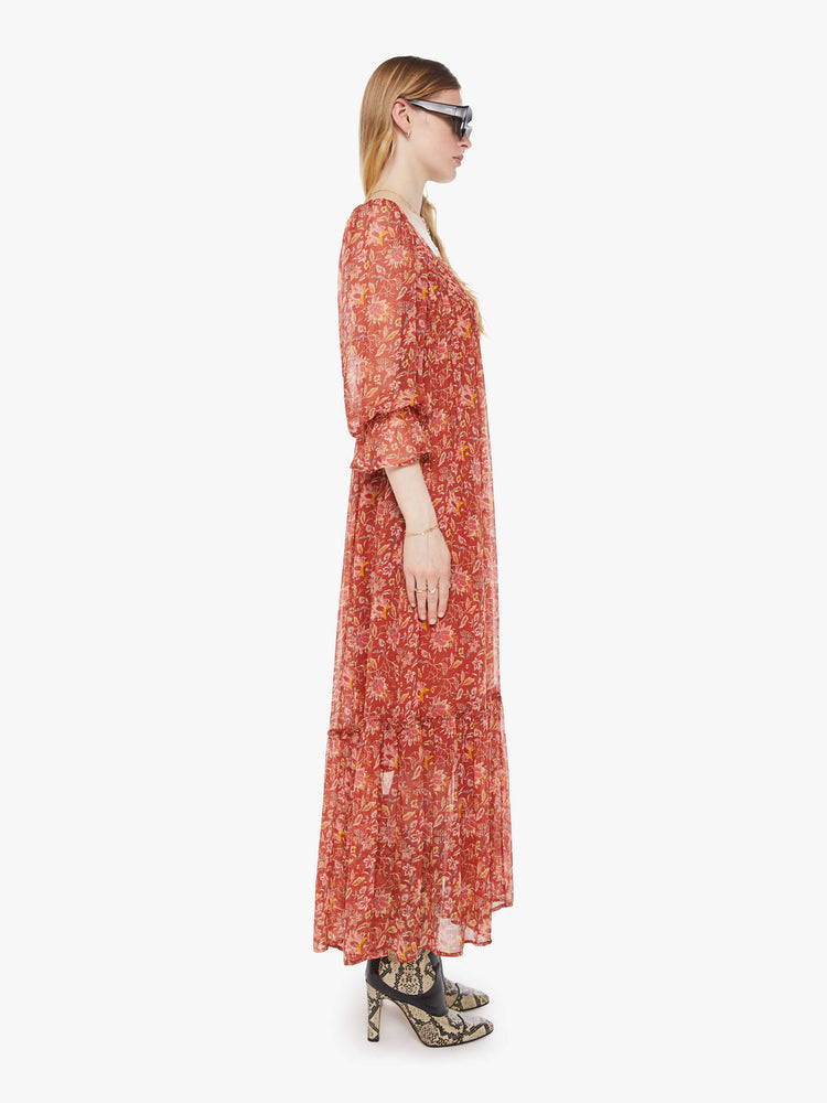Side view of a woman in a burnt orange with a warm-toned floral print dress with a square neck, 3/4-length sleeves, ruffled details and a loose, flowy fit.