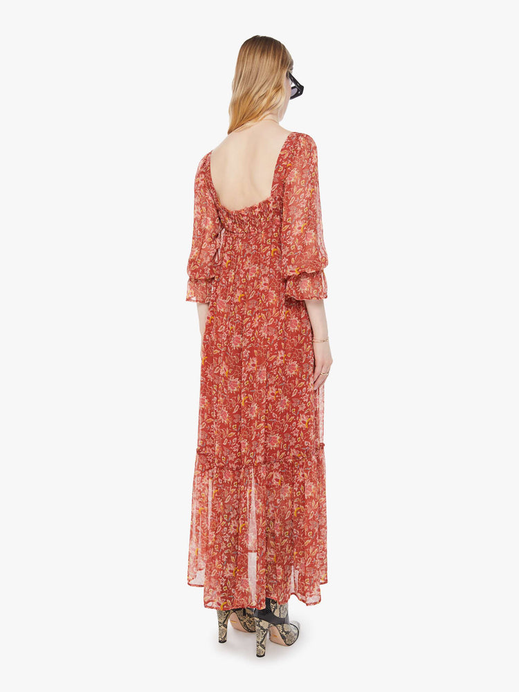 Back view of a woman in a burnt orange with a warm-toned floral print dress with a square neck, 3/4-length sleeves, ruffled details and a loose, flowy fit.