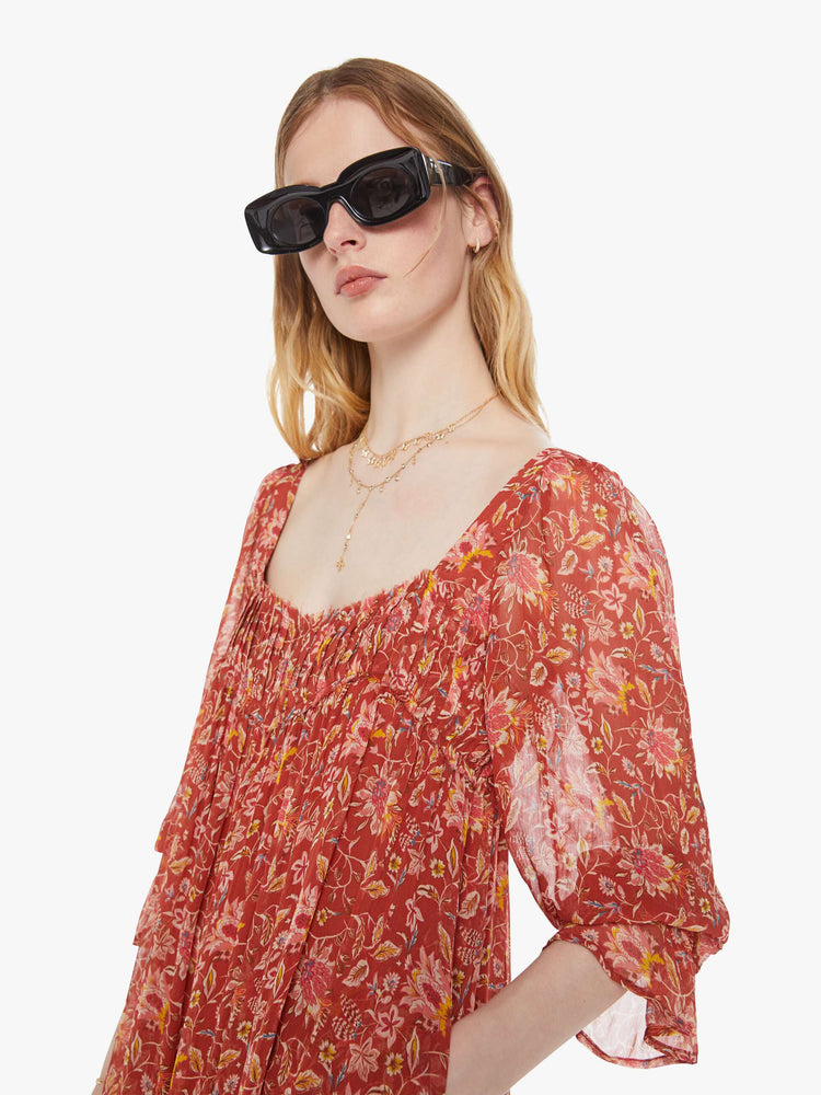 Close up view of a woman in a burnt orange with a warm-toned floral print dress with a square neck, 3/4-length sleeves, ruffled details and a loose, flowy fit.
