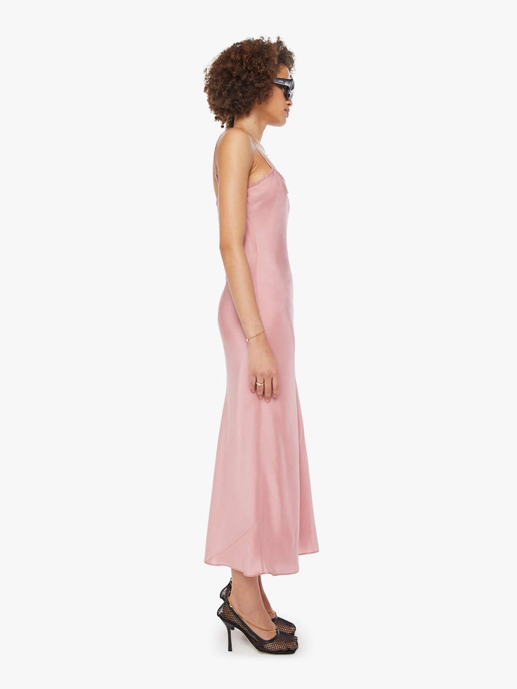 Side view of a woman  in pink dress with thin spaghetti straps, a curved neck, narrow fit and a flowy ankle-length hem.