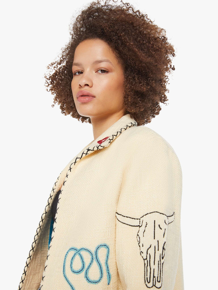 Close up view of a woman in a cream jacket with beaded motifs and slightly dropped shoulders, front patch pockets and a tailored fit that hugs the body like a blazer.
